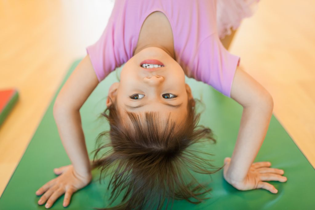 A happy child in a pink shirt performing gymnastic bridge pose on a green mat indoors.