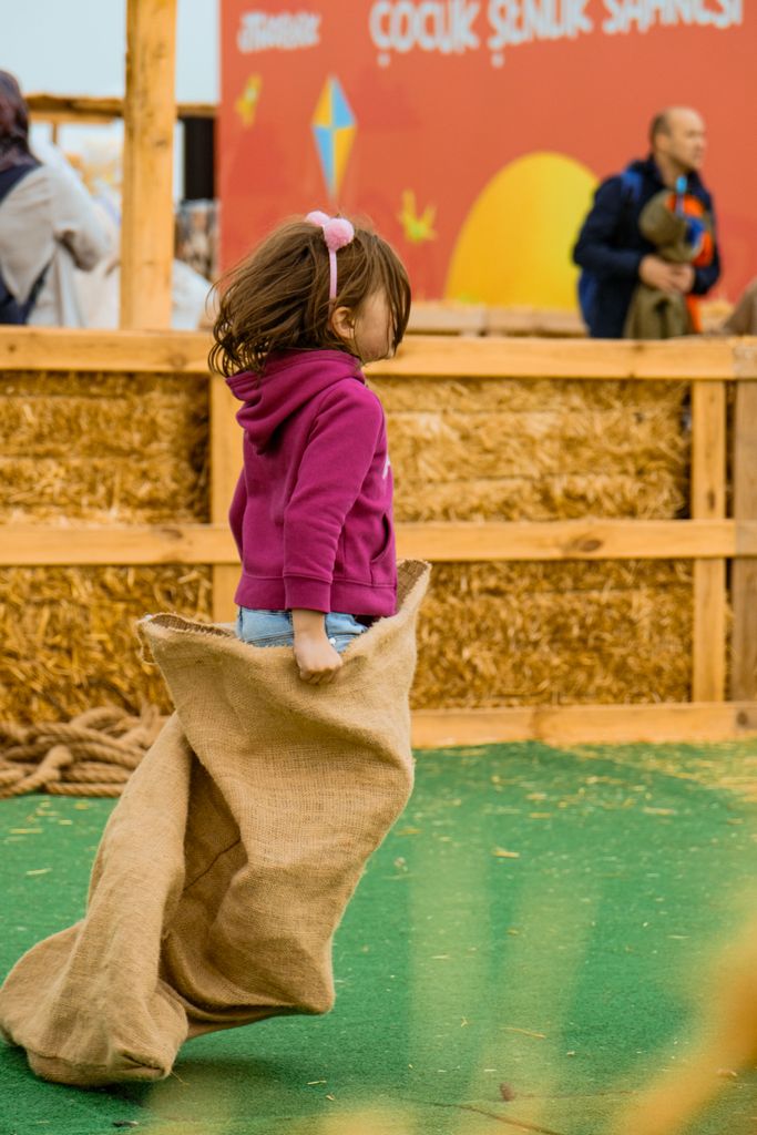 A young girl participates in a sack race during an outdoor event with hay bales and colorful signage.
