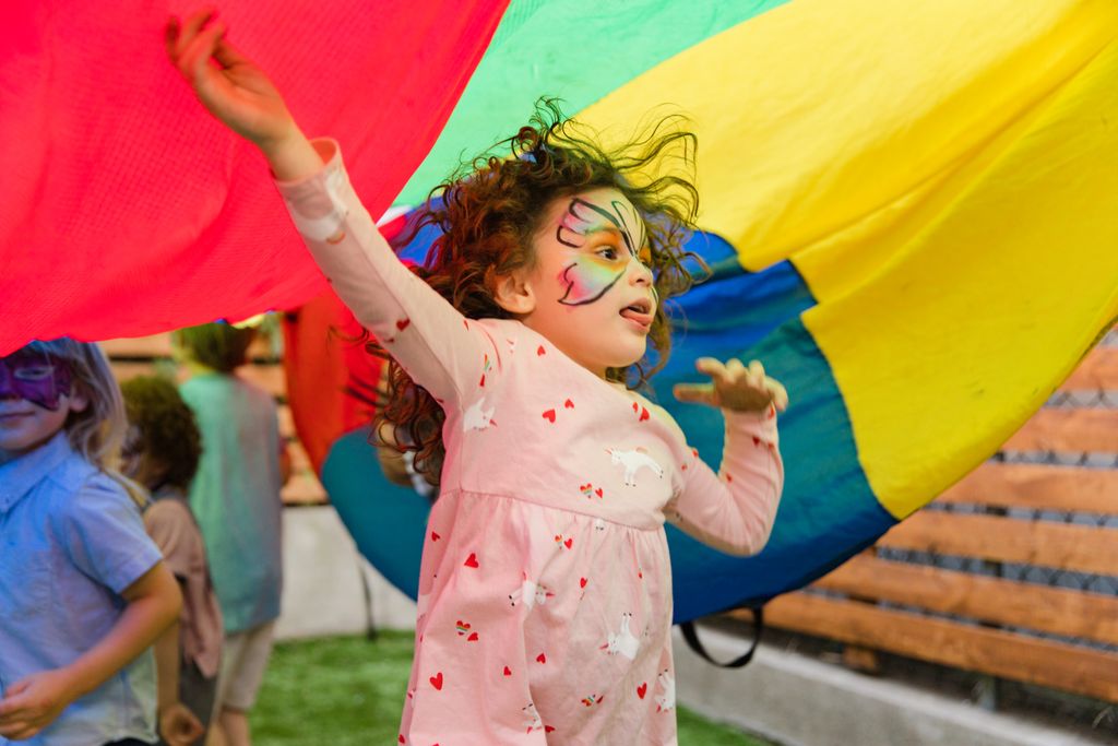 Children enjoying a lively outdoor party under vibrant parachute play, face painted with colorful designs.