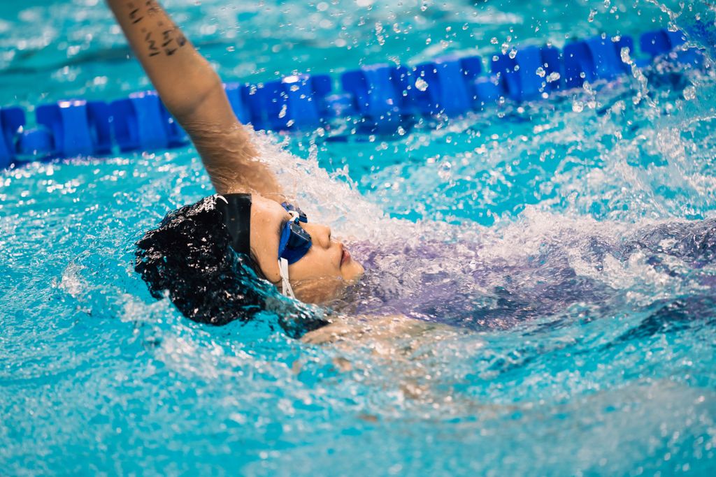 Swimmer doing backstroke in pool with blue goggles during a competitive event.