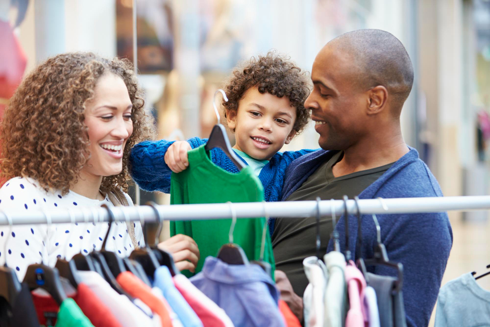 A man holding a child while a woman smiles as they pull clothes off a rack in a store.